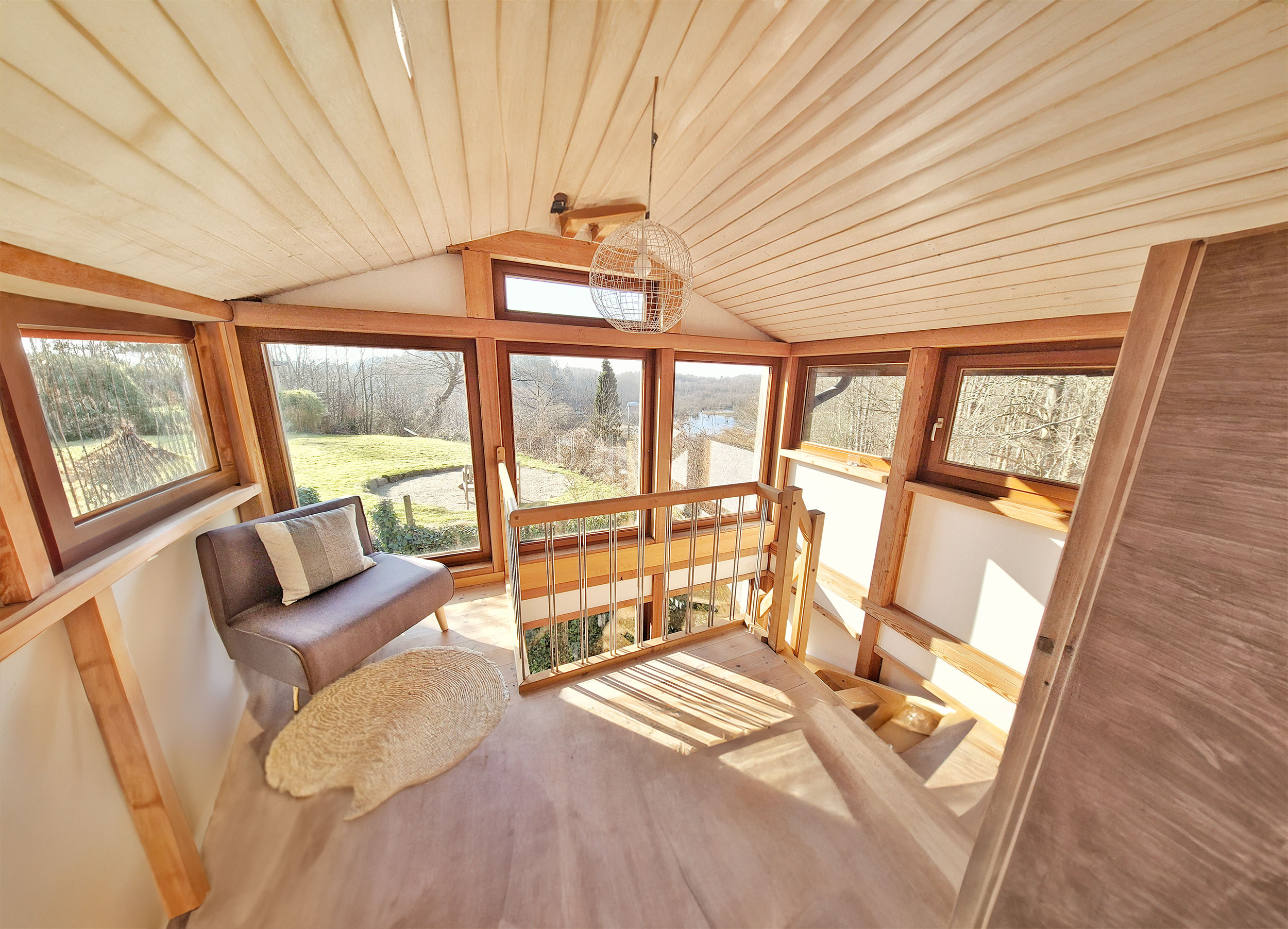 Gemütliches Wohnzimmer mit heller Holzverkleidung, grünem Ausblick und einem grauen Sofa vor großen Fenstern.
