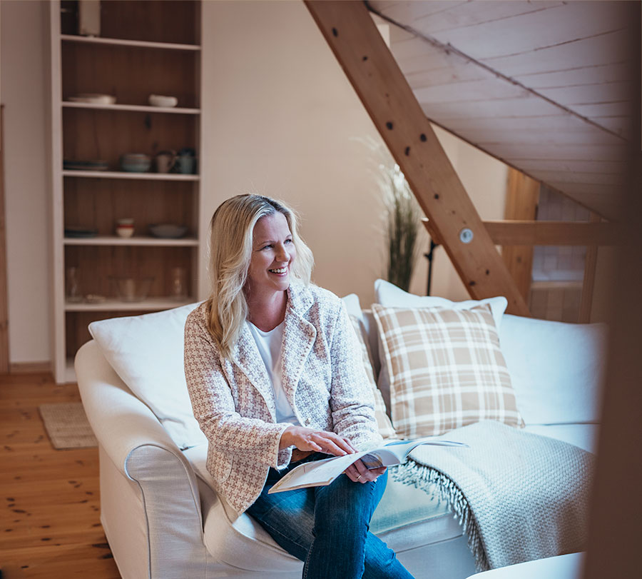Lächelnde Immobilienmaklerin mit blonden Haaren auf einem weißem Sofa, sie hält eine Broschüre in ihren Händen. Sie befindet sich in einem hellen Dachgeschoss mit natürlichem Licht.