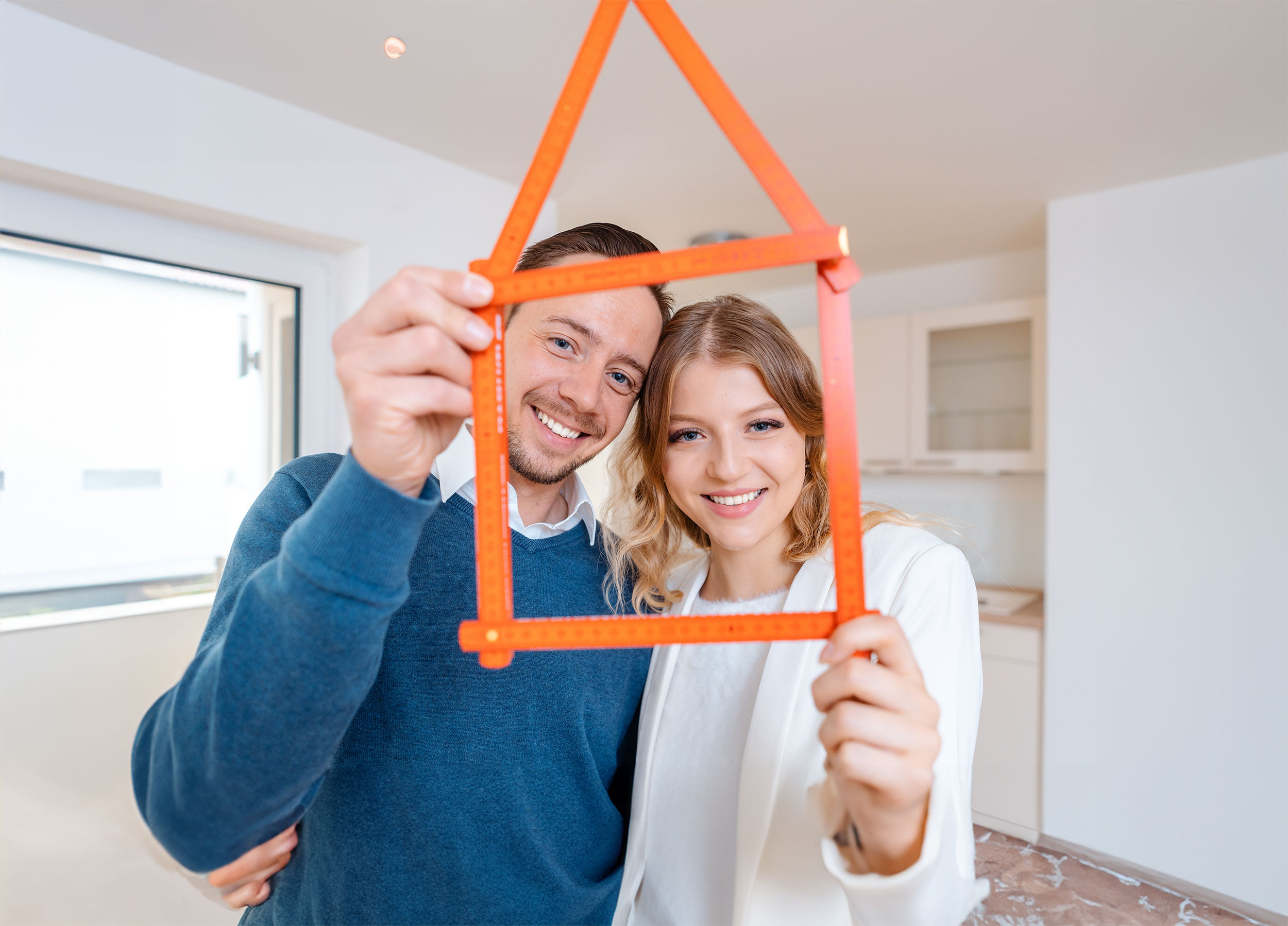 Lächelndes junges Paar hält symbolisch ein kleines Haus in den Händen in einem modernen Neubauraum.