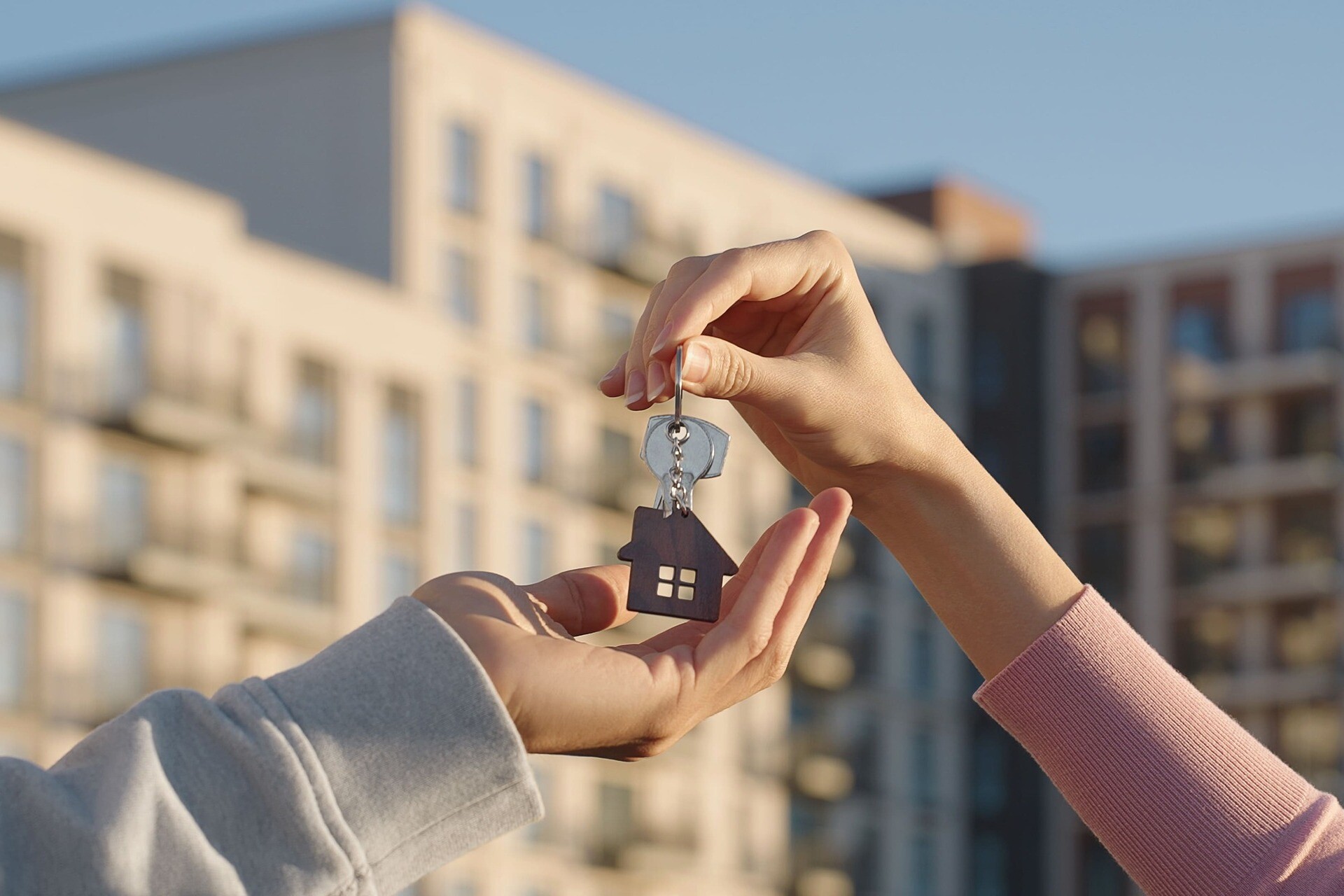 Nahaufnahme einer Immobilienbesitzerin, die einem jungen Mann im Freien vor dem Hintergrund eines modernen Wohnhauses einen Hausschlüssel mit einem Schlüsselanhänger in Form eines Hauses überreicht.