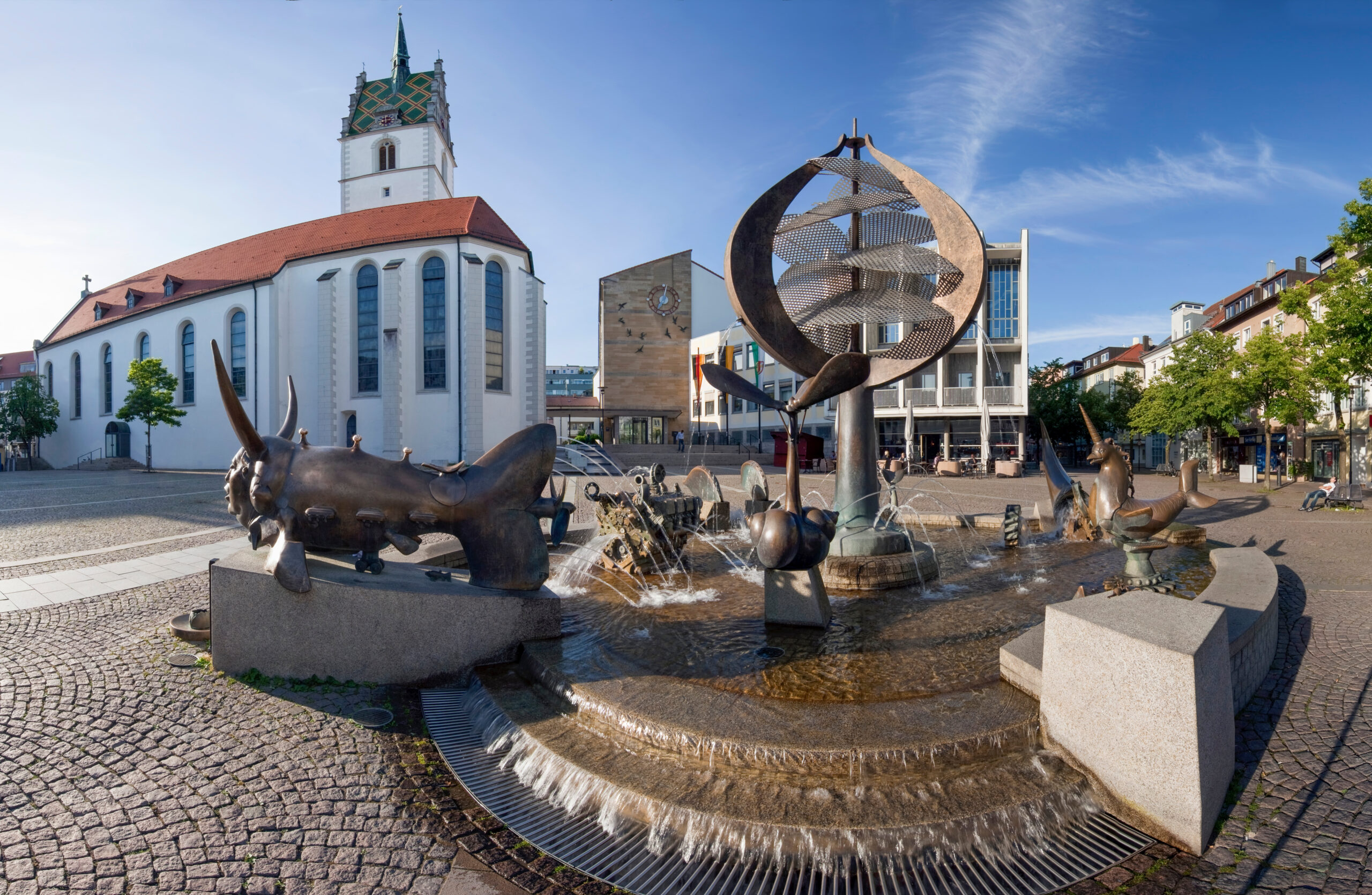 Brunnen mit Tierfiguren, Kirche und moderne Gebäude im Hintergrund.