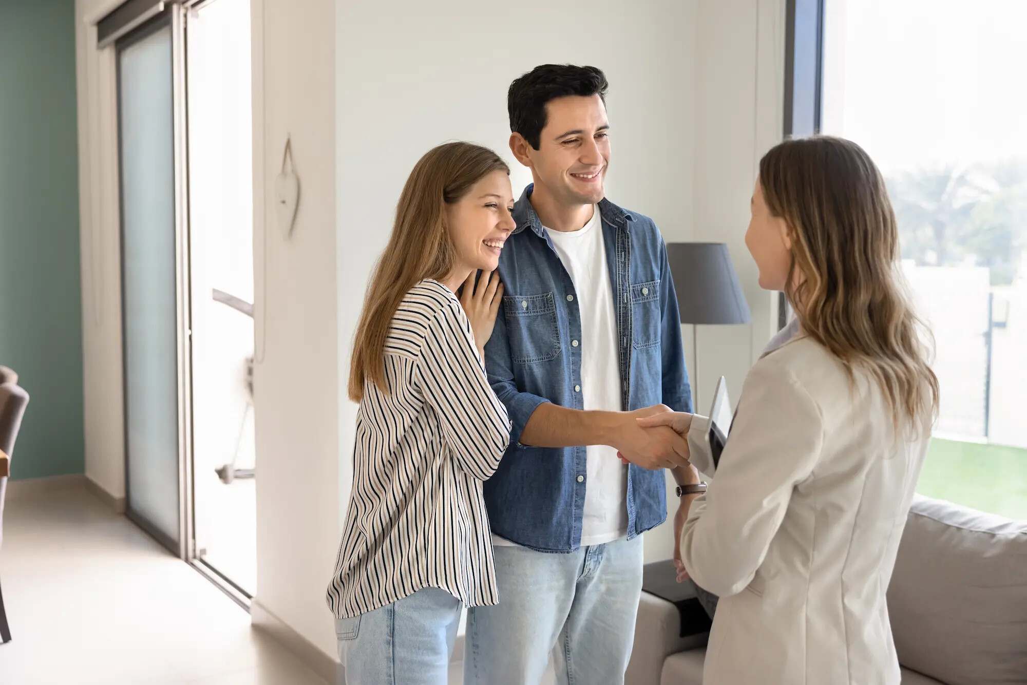 Glückliches junges Paar besichtigt moderne Wohnung und begrüßt Immobilienmaklerin mit Handschlag bei erfolgreicher Hausbesichtigung