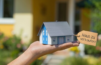 Person hält Miniaturhaus mit Verkauf-Schild auf der Hand