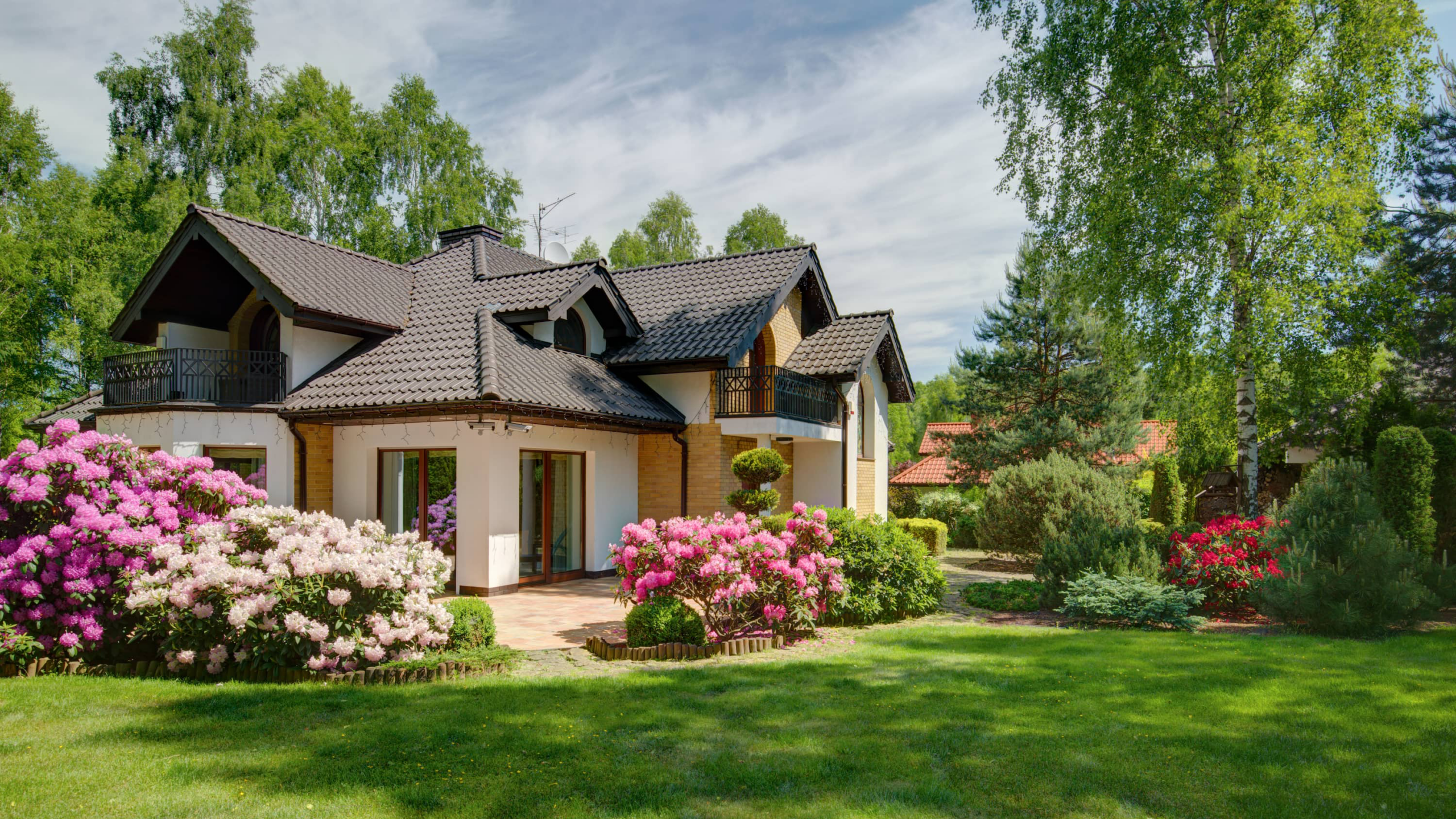 Haus mit dunklem Dach, bunte Büsche und grüne Bäume bei Sonne.