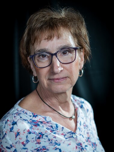Professionelles Business-Porträt einer älteren Frau mit Brille und kurzen braunen Haaren. Sie trägt eine moderne Bluse mit abstraktem Muster und dezente Schmuckstücke. Das Studiofoto mit dunklem Hintergrund hebt ihre freundliche und selbstbewusste Ausstrahlung hervor.