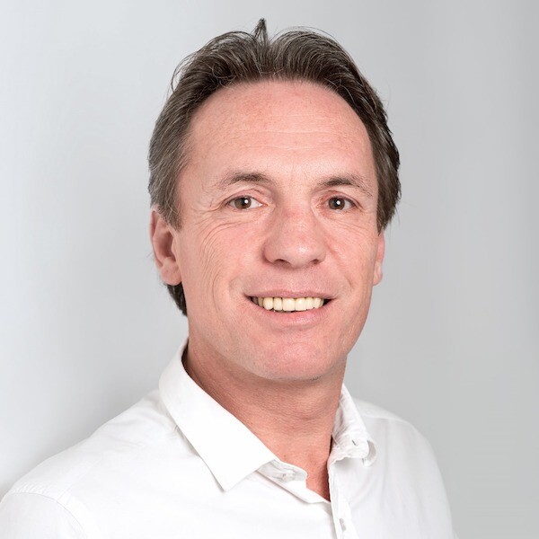 Christian Verhunz. Professionelles Bewerbungsfoto eines lächelnden, sympathischen Mannes mittleren Alters mit braunen Augen und dunklen, leicht graumelierten Haaren, der ein weißes Hemd vor einem neutralen grauen Hintergrund trägt.
