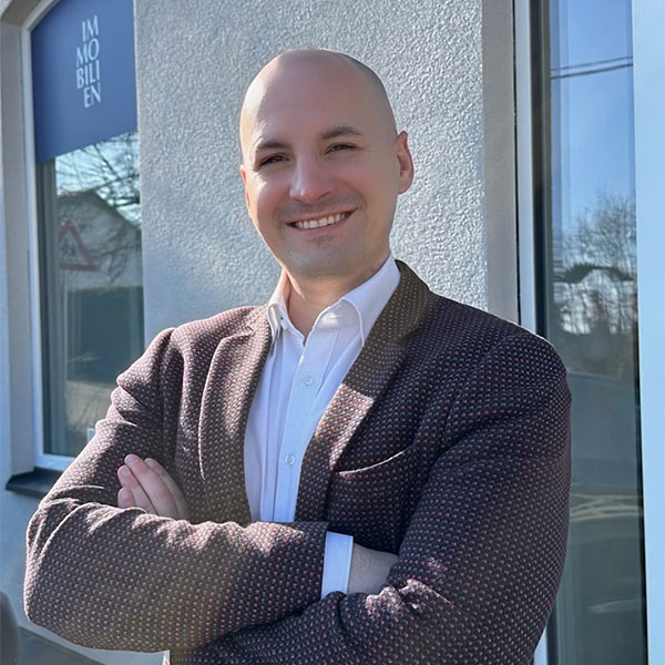 Marc-André Schenner. Professioneller Immobilienmakler lächelt mit verschränkten Armen vor einem Immobilienbüro in bei sonnigem Wetter.