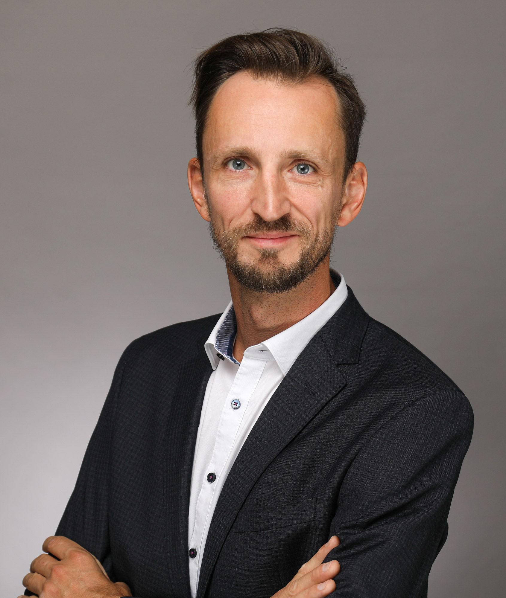 Michael Sax. Professionelles Bewerbungsfoto eines lächelnden Mannes mit Bart, blauem Hemd und Sakko vor grauem Hintergrund.