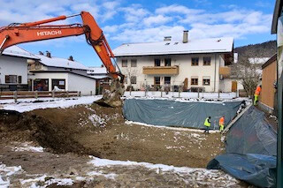 Bagger gräbt nahe Häusern; Arbeiter im Schnee in Sicherheitskleidung.