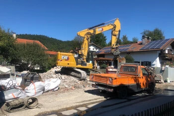 Gelber Bagger auf Schutt, oranger Lkw, Häuser mit Solarpanels.