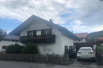 Weißes Haus, dunkles Dach, Balkon, zwei Autos, Holzzaun, Wolken.