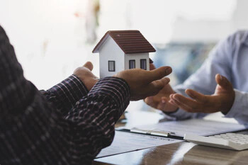 Person hält Modellhaus über Tisch mit Dokumenten und Laptop.
