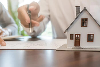 Hand mit Stift bei Hausplänen und Modellhaus auf Schreibtisch.