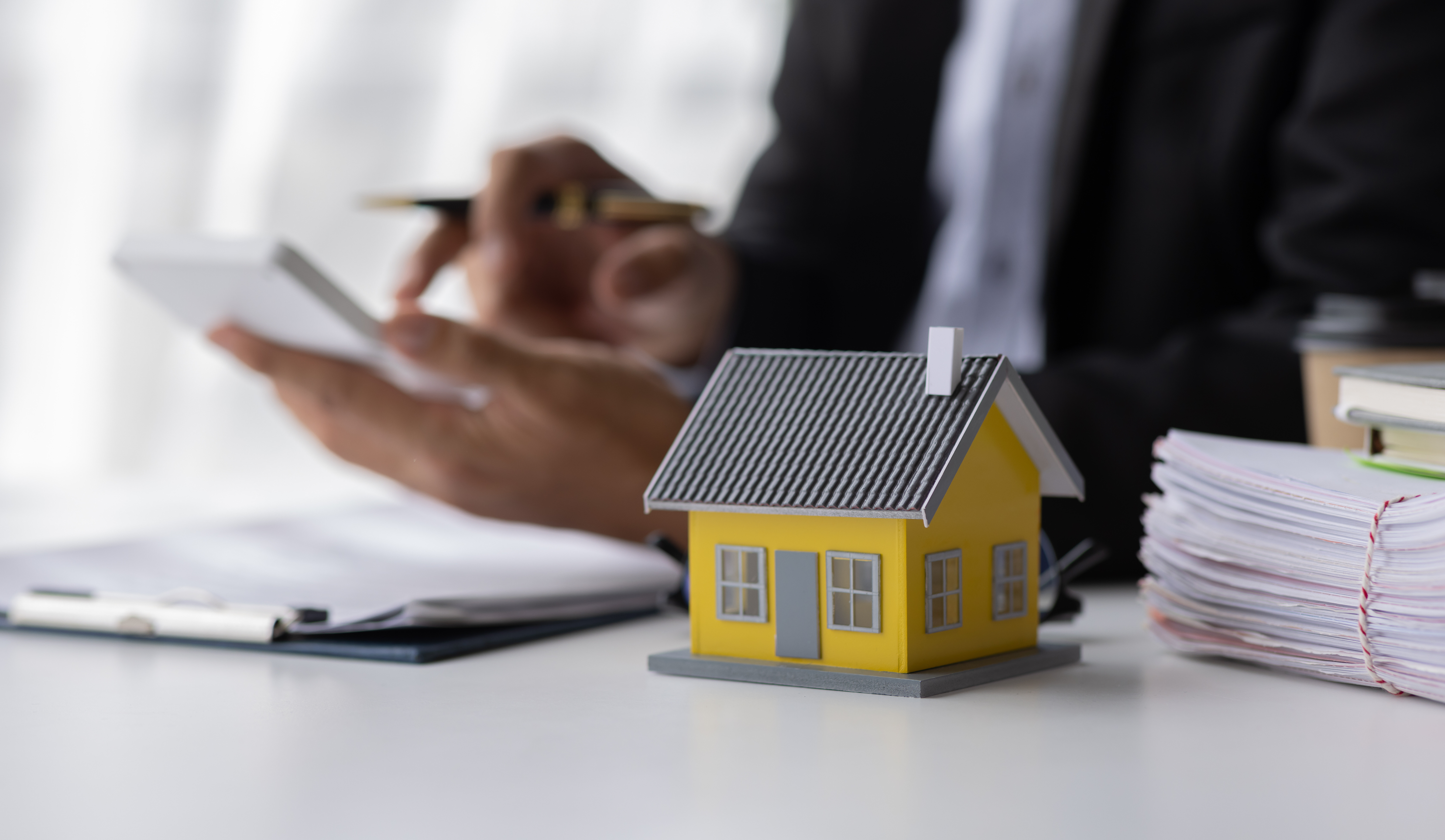 Ein gelbes Modellhaus steht auf einem Schreibtisch, im Hintergrund tippt ein Mann etwas in einen Taschenrechner