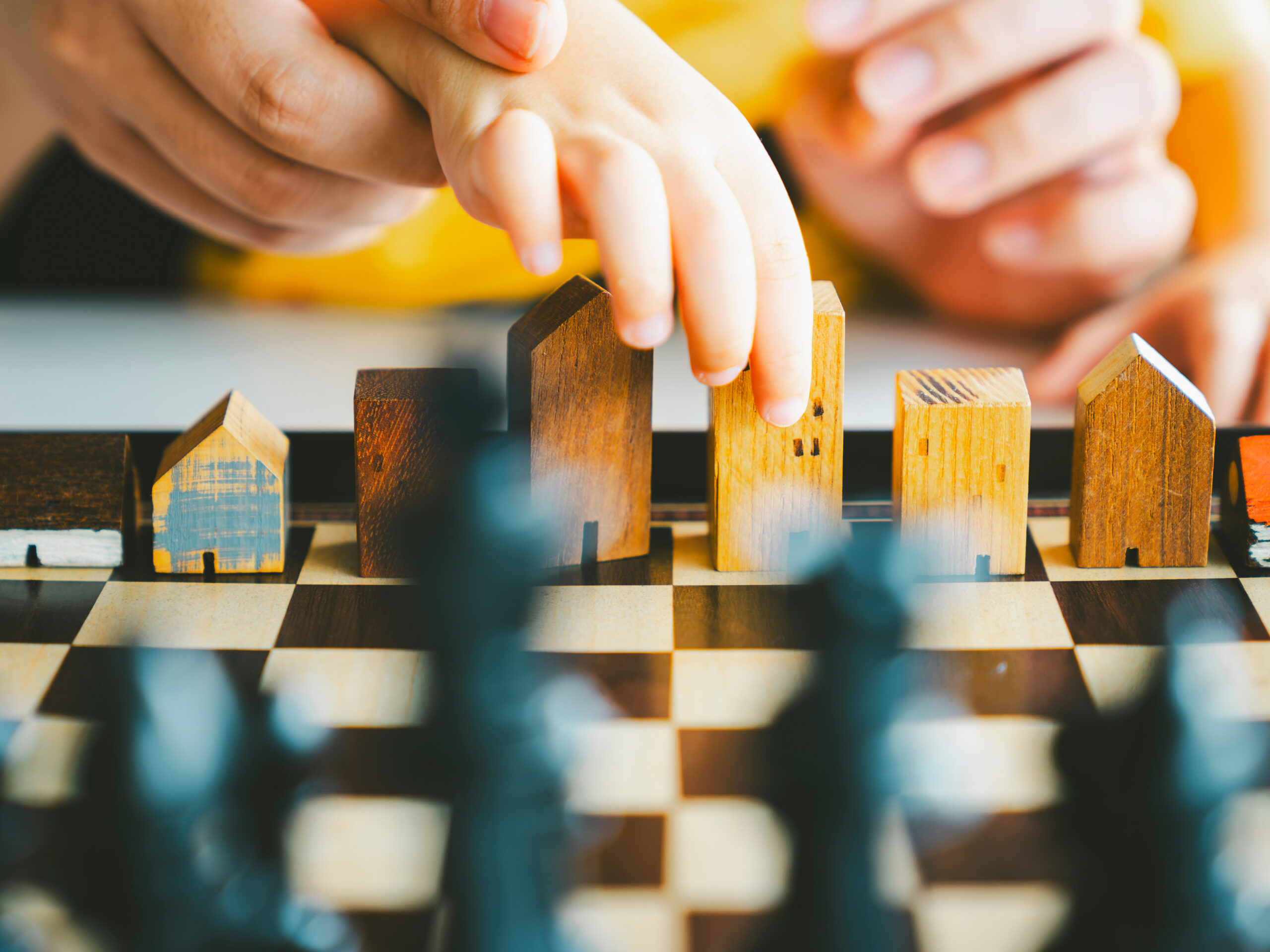 Eine Kinderhand hält ein Modellhaus, das neben anderen Modellhäusern auf einem Schachbrett steht