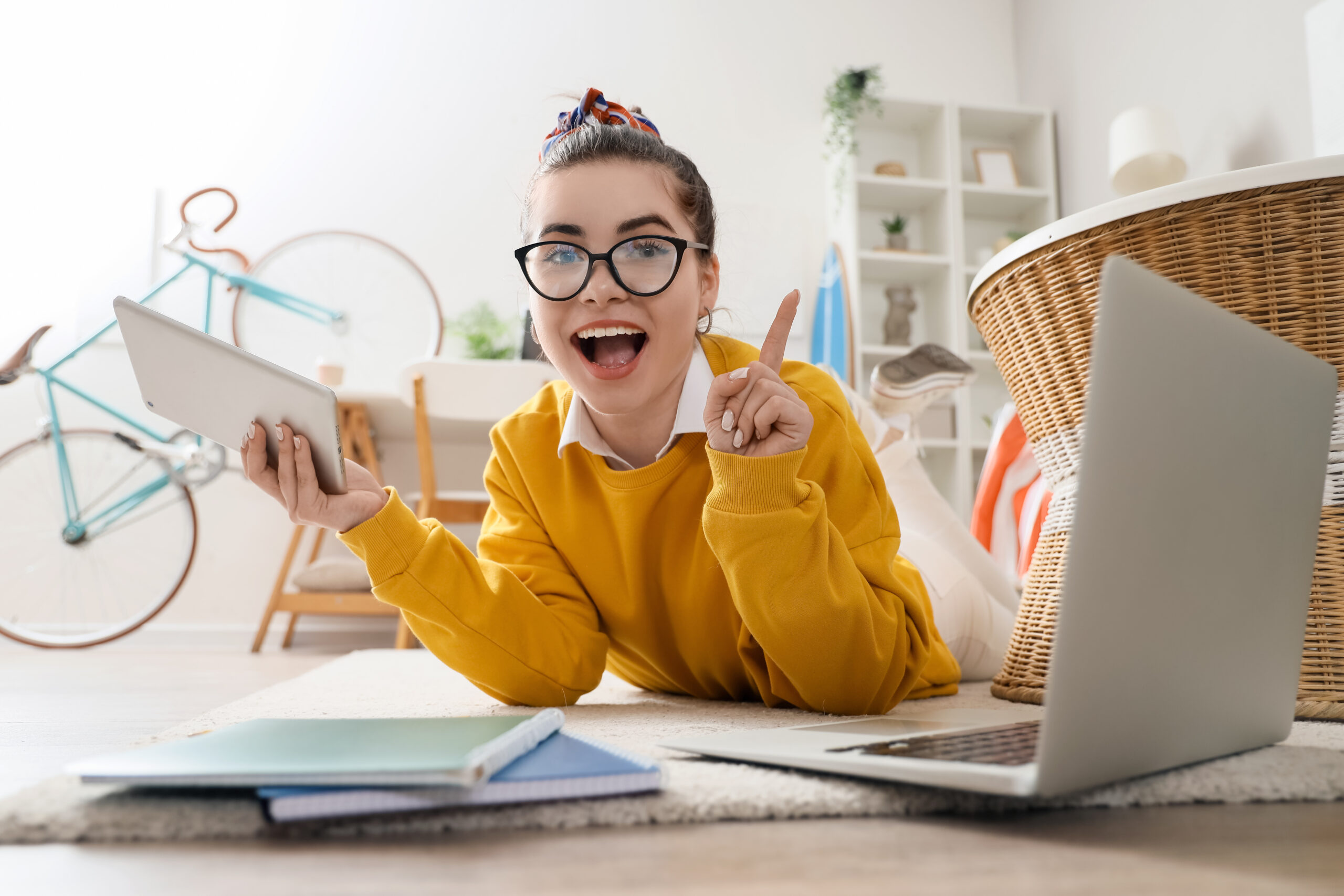 Eine junge Frau liegt auf dem Bauch auf dem Boden, neben ihr ein Laptop und Bücher. Sie schaut in die Kamera, als ob ihr eine Immobilie gefällt