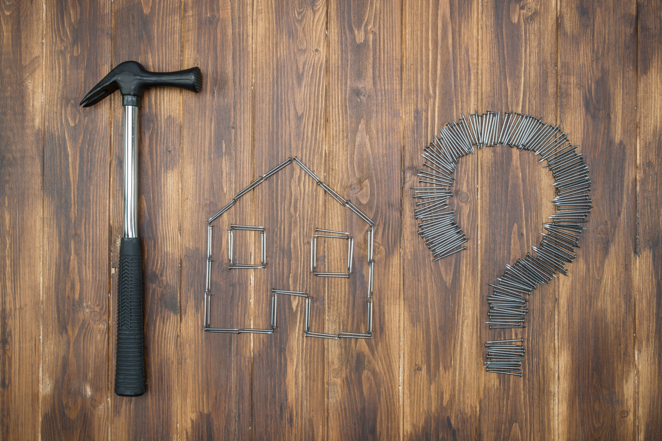 Ein Hammer liegt neben einem Haus, das aus Nägeln gelegt wurde, daneben ein Fragezeichen