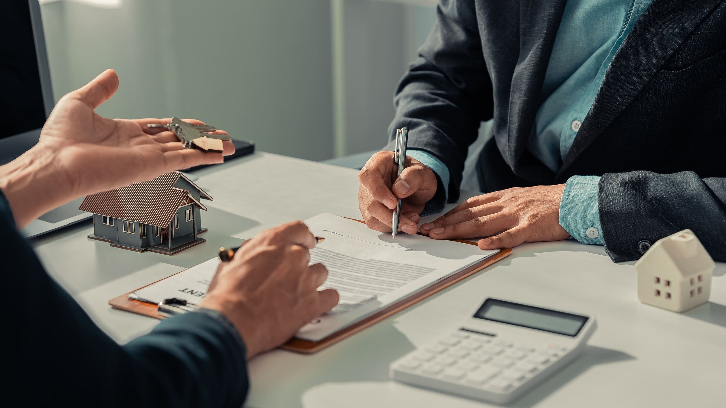 Kunde unterschreibt einen Makler-Alleinauftrag, daneben Modellhaus und Taschenrechner