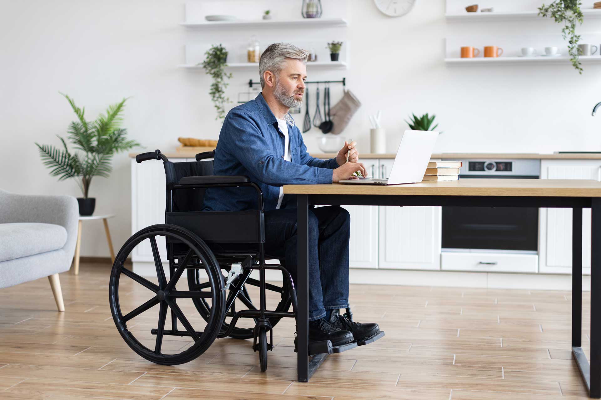 Mann im Rollstuhl sitzt mit Laptop am Küchentisch in seiner barrierefreien Wohnung