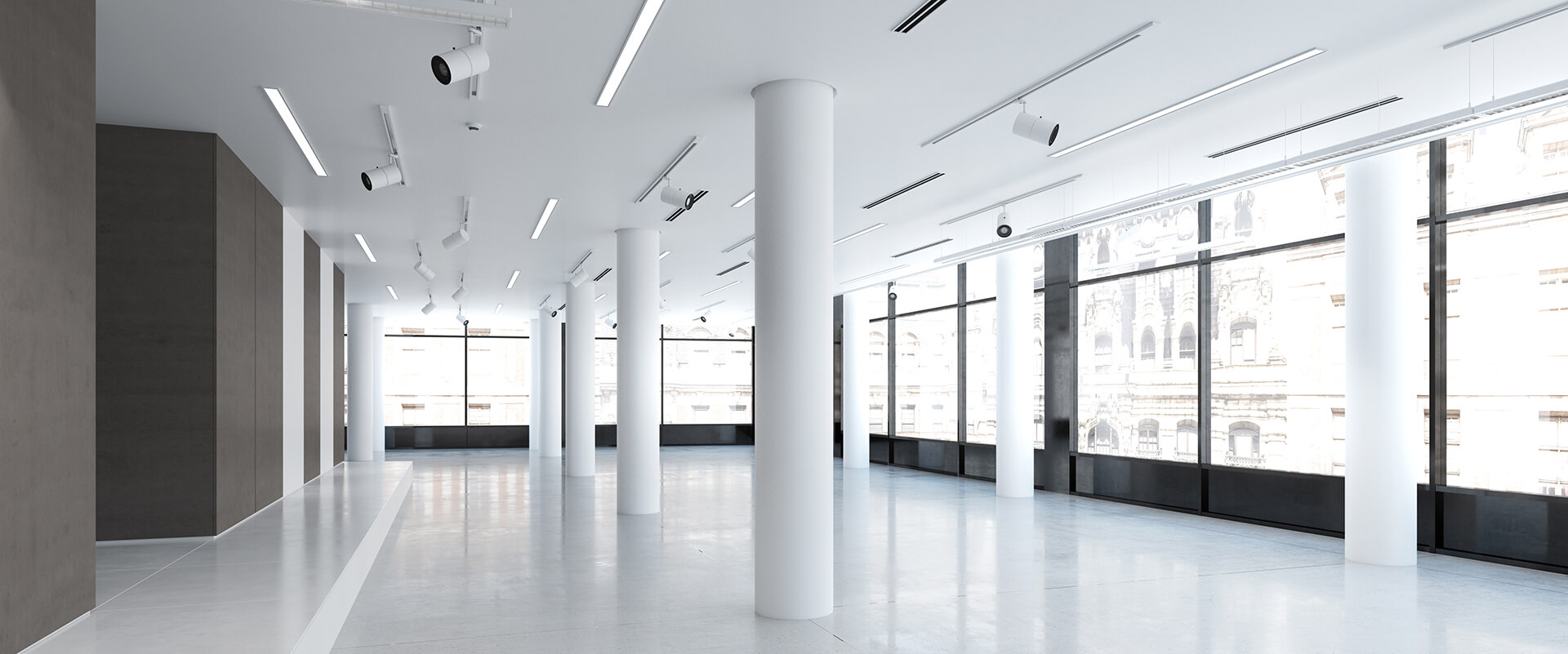 Leerer Raum mit weißen Säulen, großen Fenstern und Deckenlichtern.