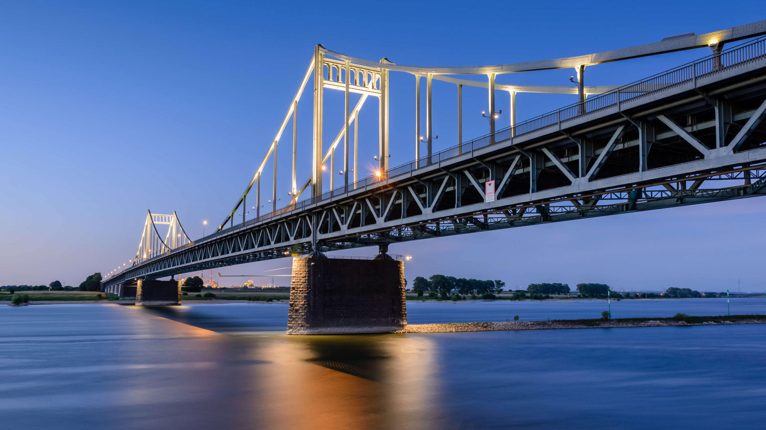 Beleuchtete Hängebrücke bei Dämmerung über ruhigem Fluss.