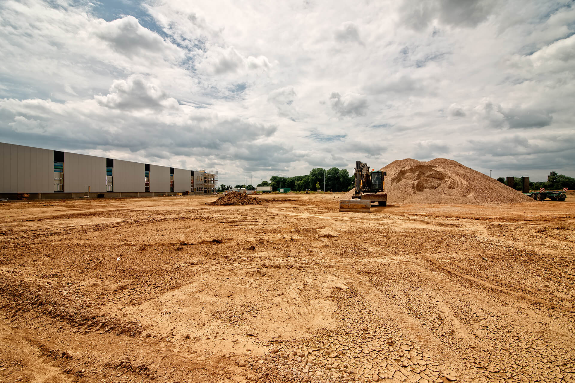 Baustelle mit Bagger, Kieshaufen und Gebäuden unter Wolken.