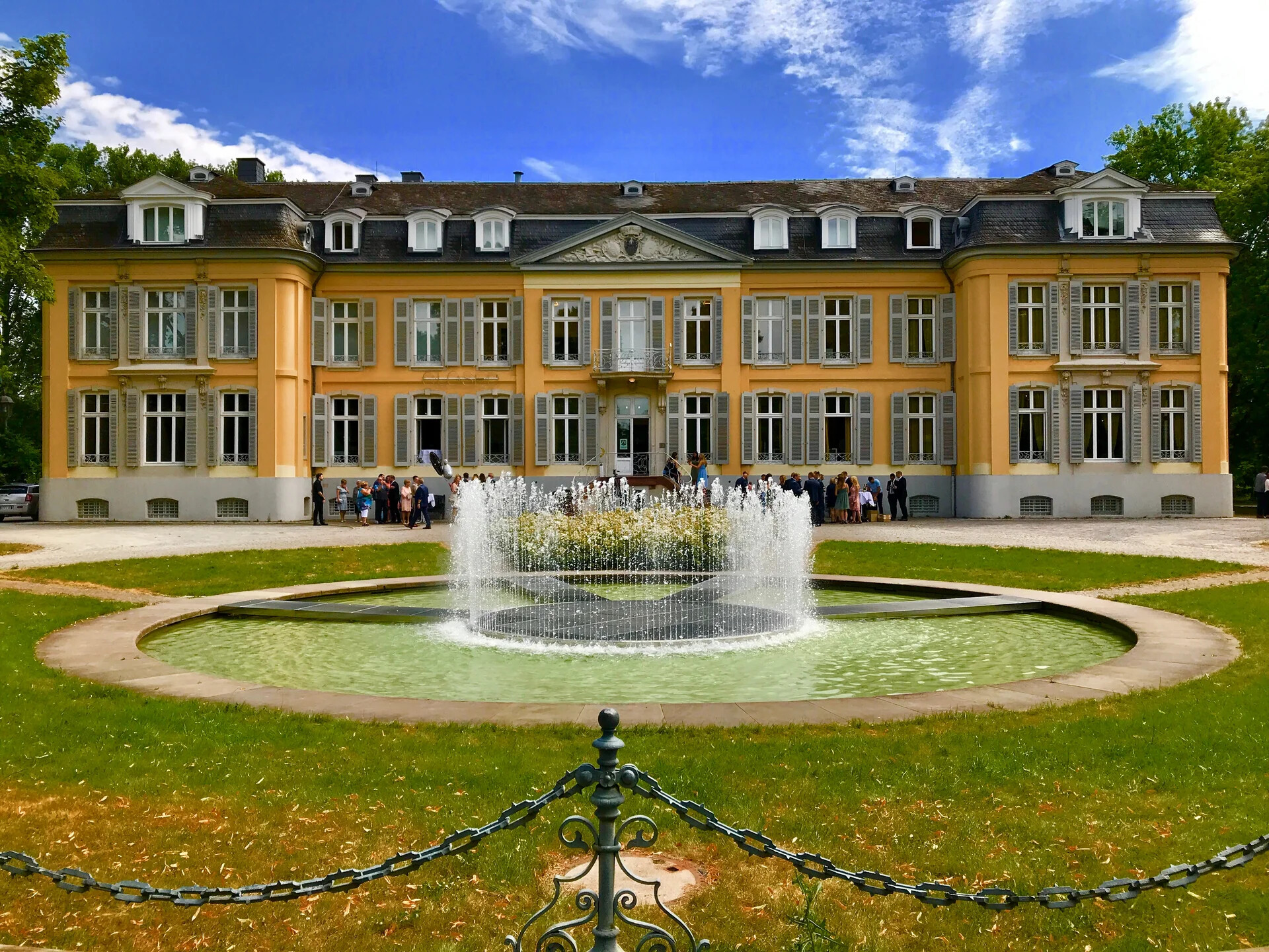 Gelbes Herrenhaus, großer Brunnen, Menschen am Eingang.