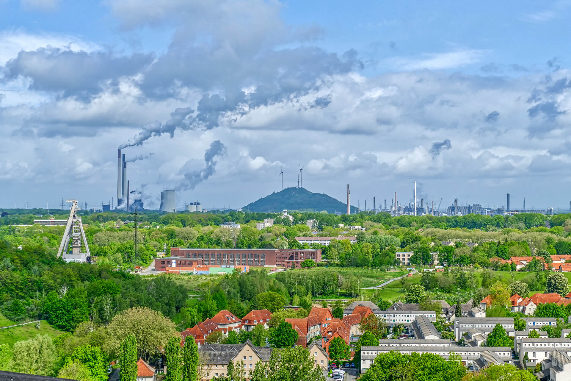 Industrieanlagen mit Rauch hinter grünem Wohngebiet und Hügel.