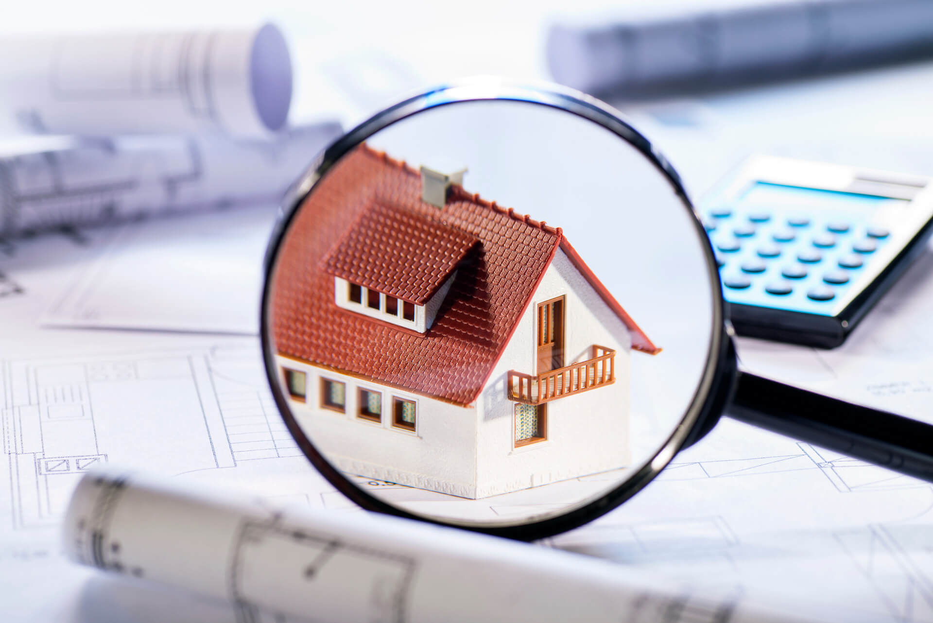 Lupe über Modellhaus, im Hintergrund Pläne und Taschenrechner.