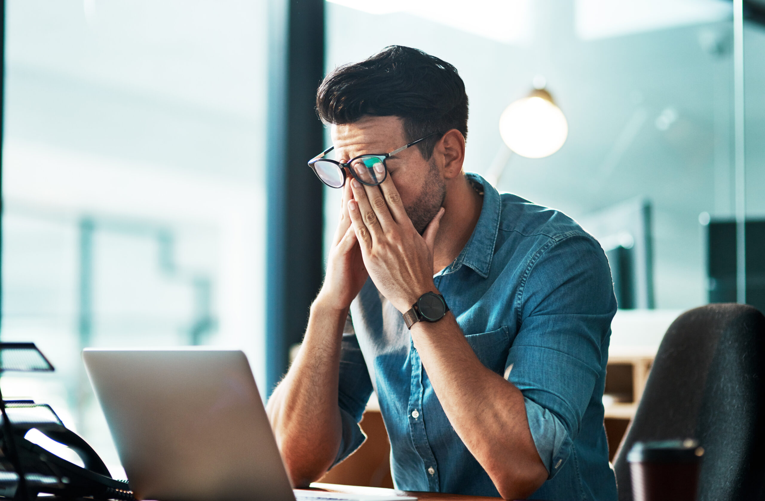 Gestresster Mann mit Brille verdeckt Gesicht am Schreibtisch, Laptop davor: Hürden beim Immobilienverkauf
