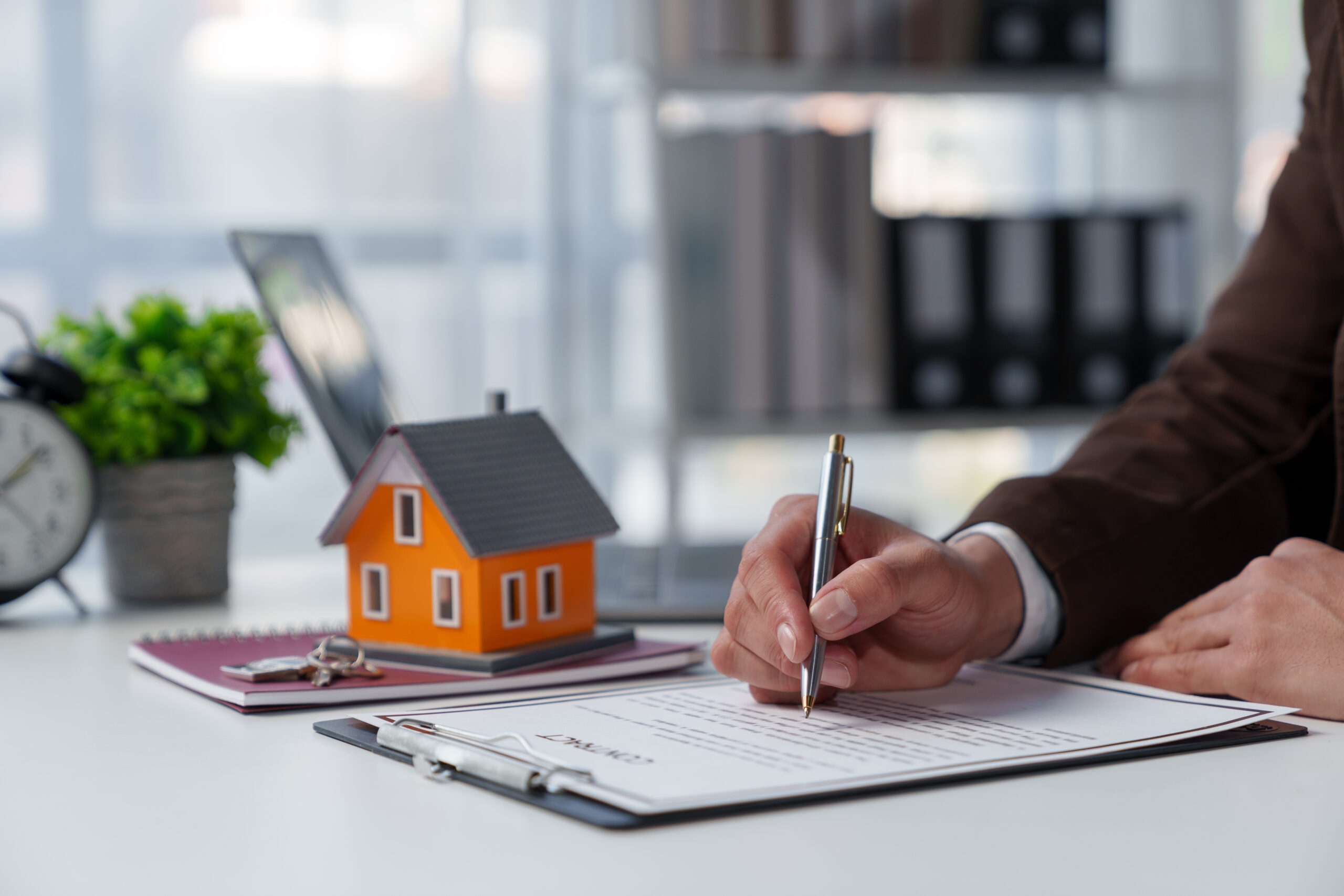 Person unterzeichnet Dokument am Schreibtisch mit Hausmodell und Büroartikeln.