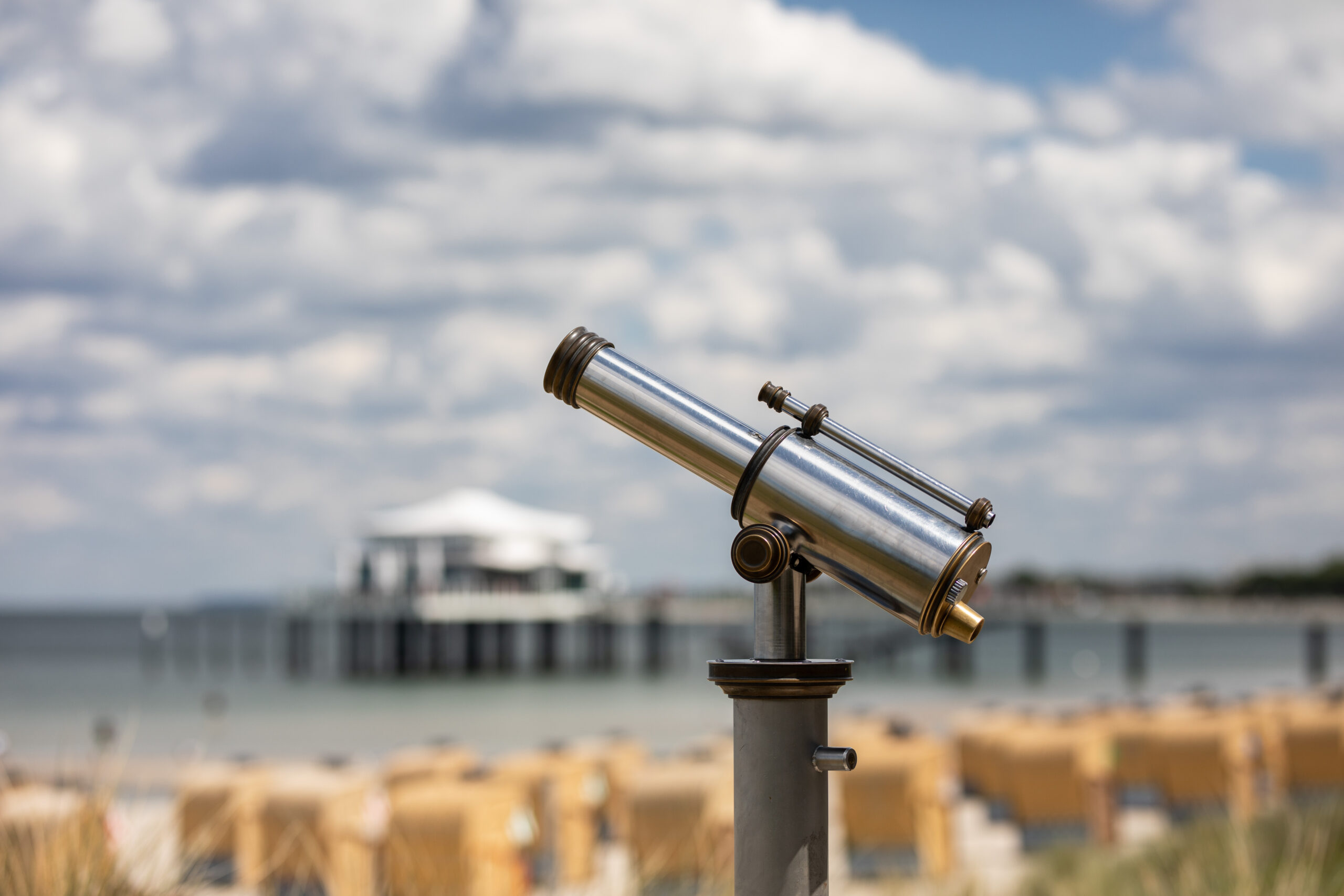 Silbernes Teleskop mit Blick auf das Meer, dahinter verschwommener Pier und Stühle.