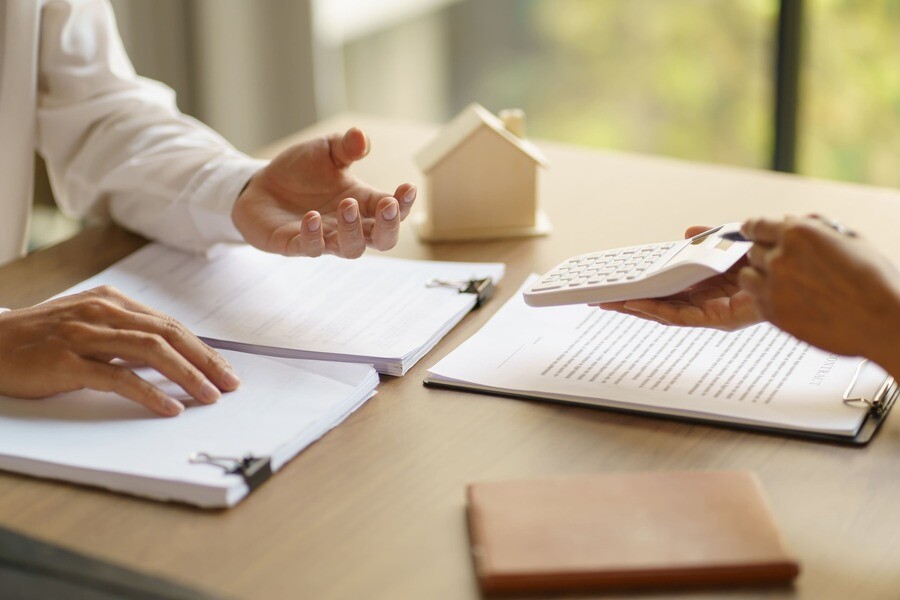 Zwei Personen mit Dokumenten, Hausmodell und Taschenrechner am Tisch