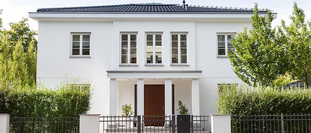 Moderne weiße Stadtvilla mit dunklem Dach und dunkler Eingangstür hinter einem Zaun.