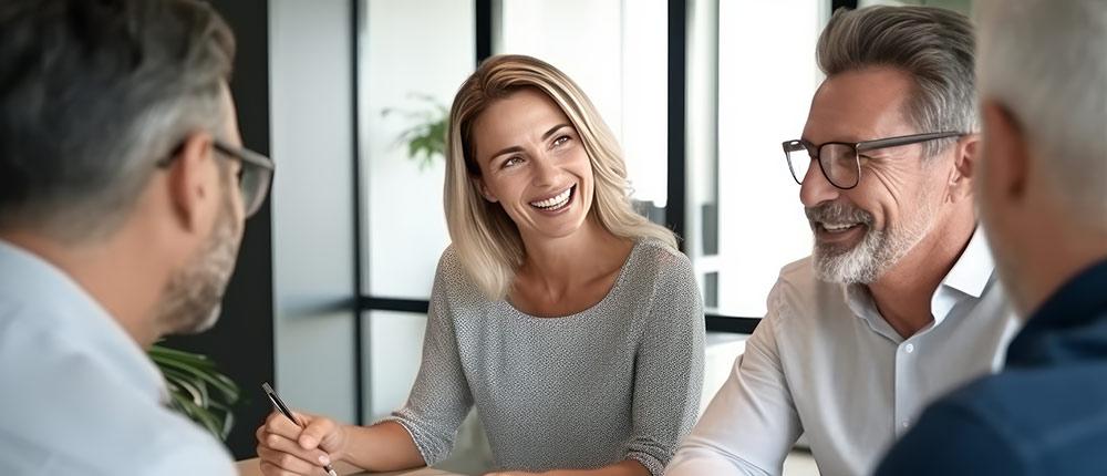 Lachendes Paar mittleren Alters sitzt mit zwei Männern in einem modernen Büro.