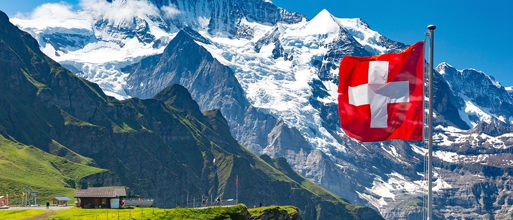 Schweizer Flagge vor schneebedeckten Alpen und grünen Bergwiesen.