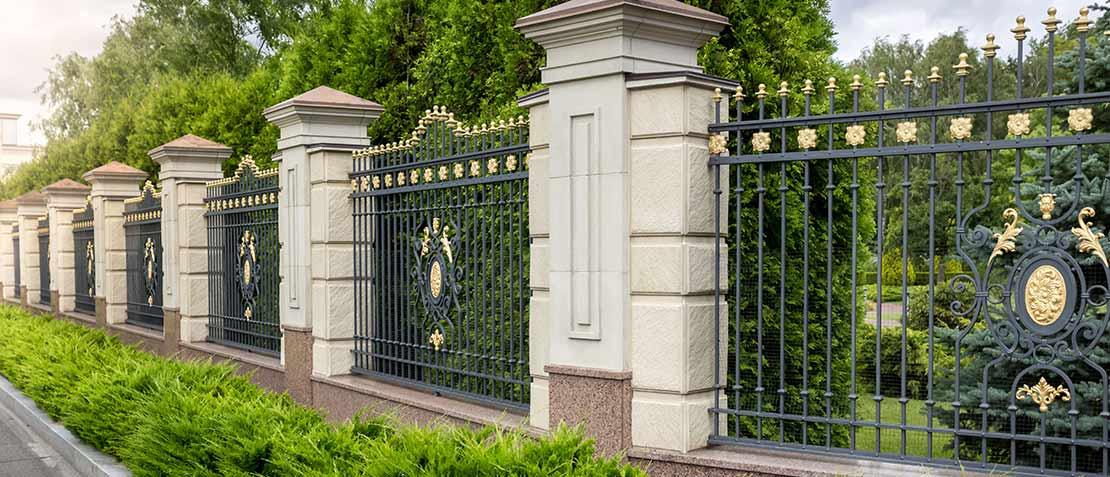 Prunkvoller Metallzaun mit verzierten Steinpfeilern vor einer grünen Hecke.