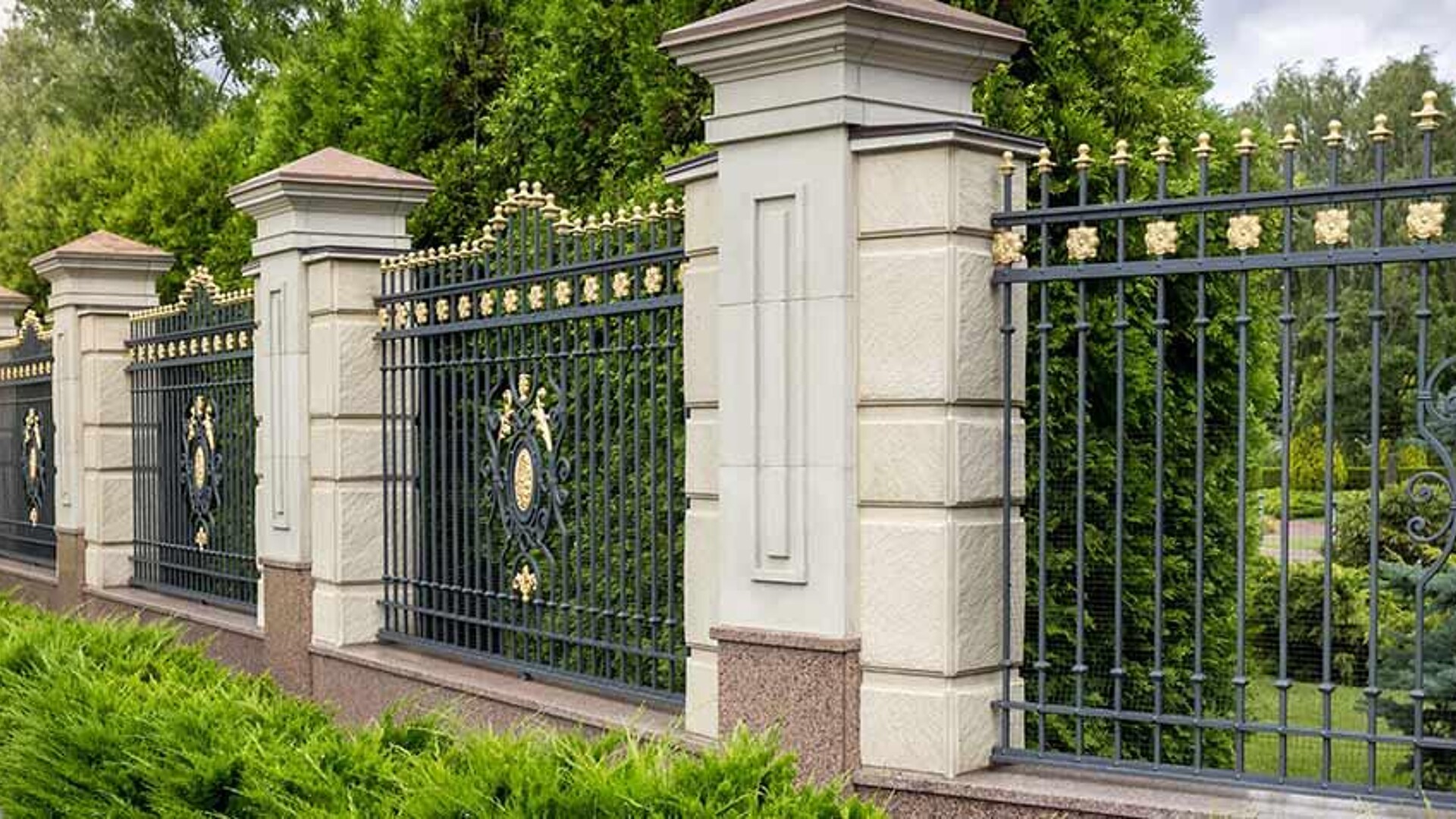 Prunkvoller Metallzaun mit verzierten Steinpfeilern vor einer grünen Hecke.