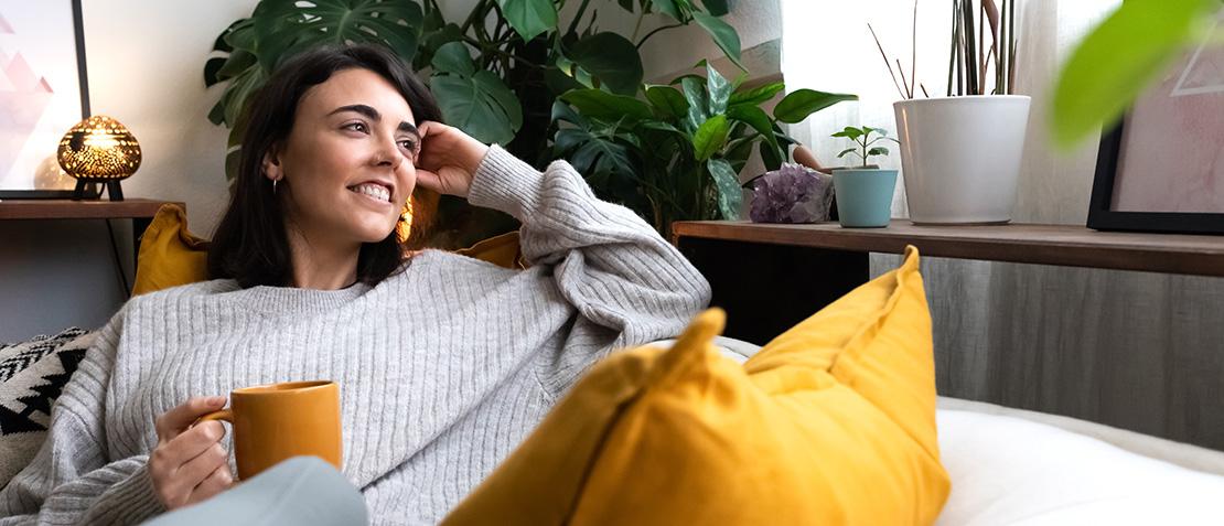 Frau sitzt lächelnd auf einer Couch mit einer Tasse in der Hand und schaut aus dem Fenster.