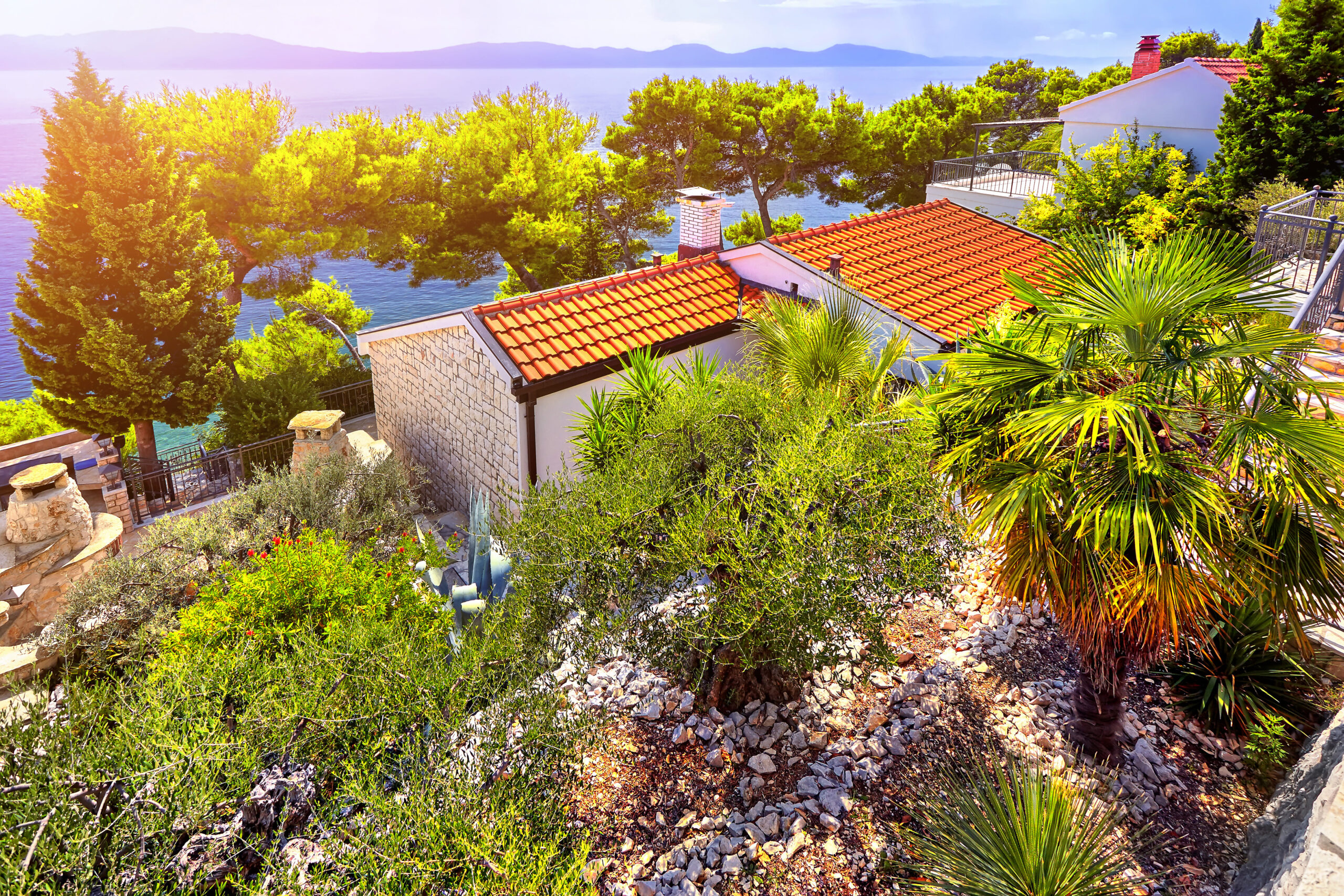 Mediterranes Haus mit roten Ziegeln umgeben von Palmen und Pinien mit Blick auf das blaue Meer.