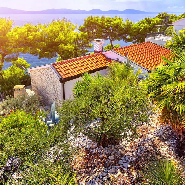 Mediterranes Haus mit roten Ziegeln umgeben von Palmen und Pinien mit Blick auf das blaue Meer.