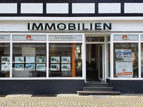 Außenansicht des Maklerbüros "Am Kirchplatz Immobilien" mit Immobilienaushängen in der Auslage.