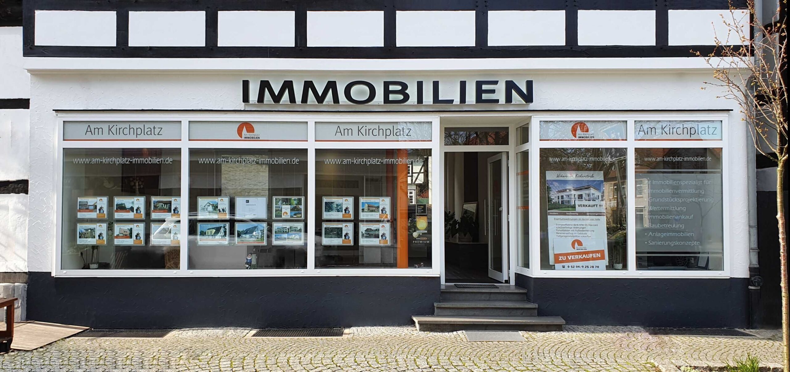 Außenansicht des Maklerbüros "Am Kirchplatz Immobilien" mit Immobilienaushängen in der Auslage.