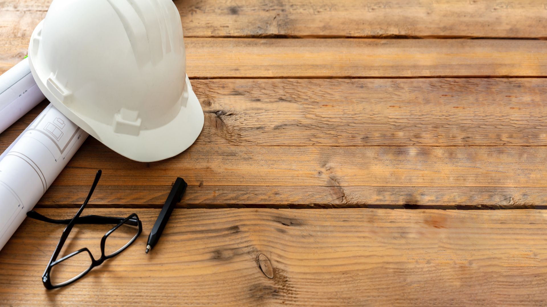 Schutzhelm, Unterlagen, Stift und Brille liegen auf einer Holzplatte als Symbol für Neubauprojekte