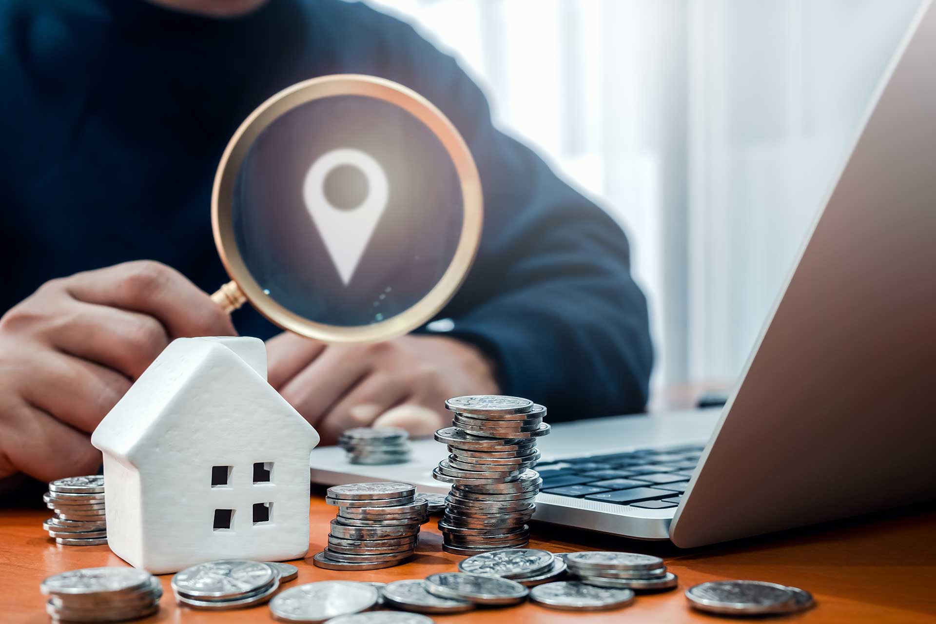 Person am Schreibtisch mit einem Modellhaus, Münzen und Laptop als Symbol für die Immobilienbewertung für den Verkauf