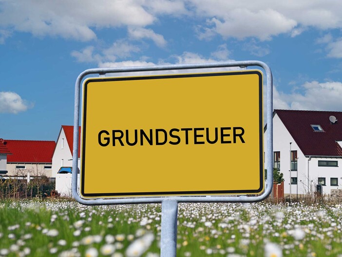 Ortsschild mit der Aufschrift 'Grundsteuer', im Hintergrund ein Wohngebiet