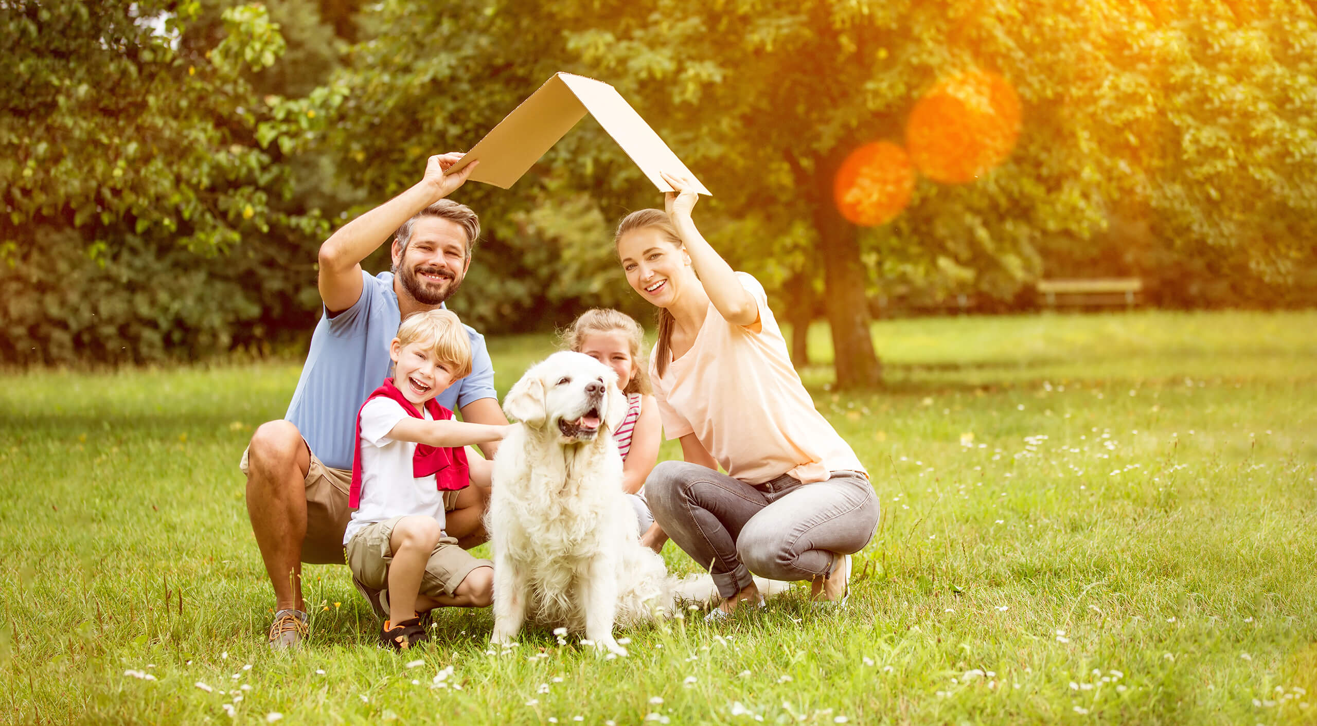 Familie mit zwei Kindern und Hund sitzt auf Gras, hält Pappdach.