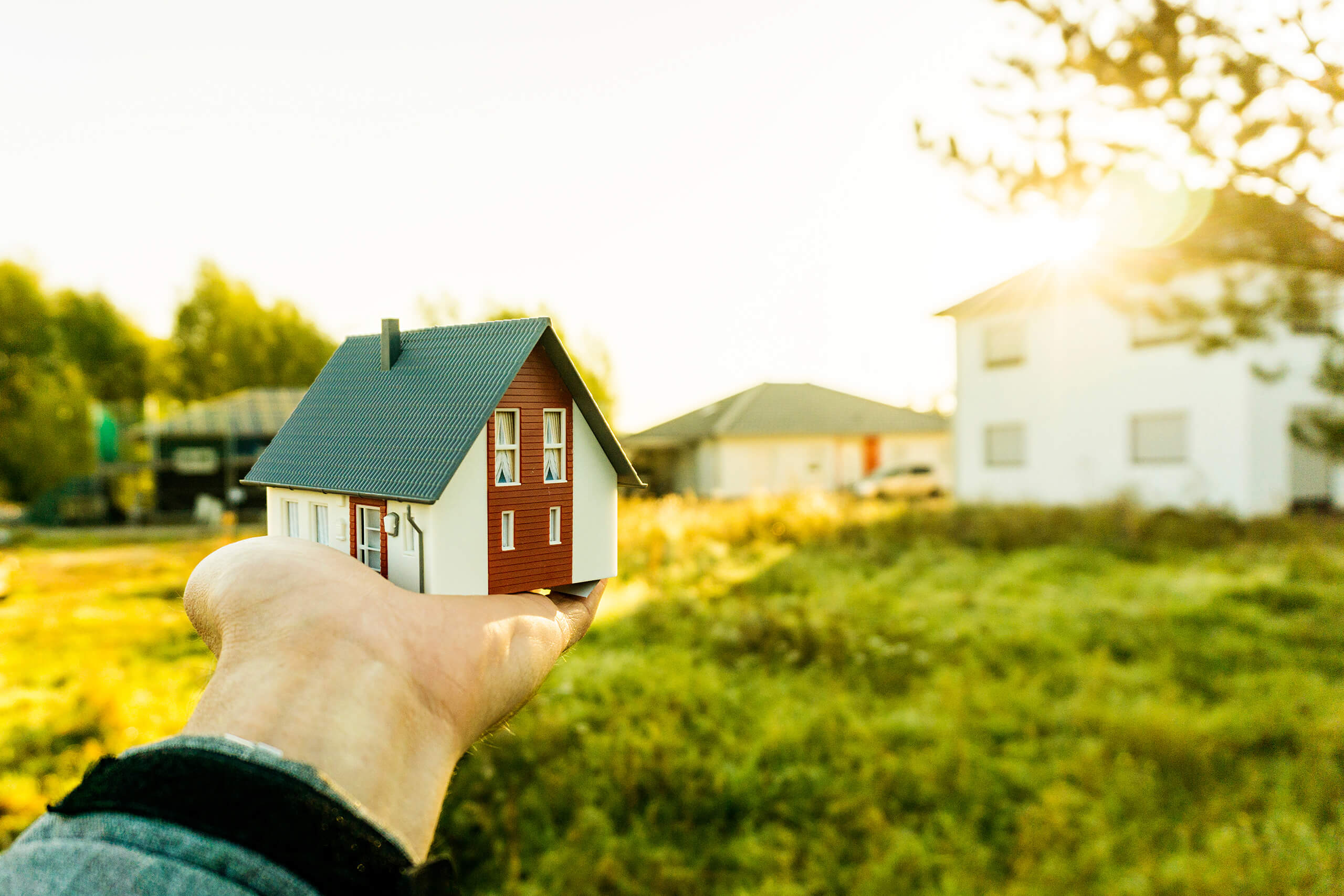 Hand hält Miniaturhaus draußen, echte Häuser im Hintergrund.