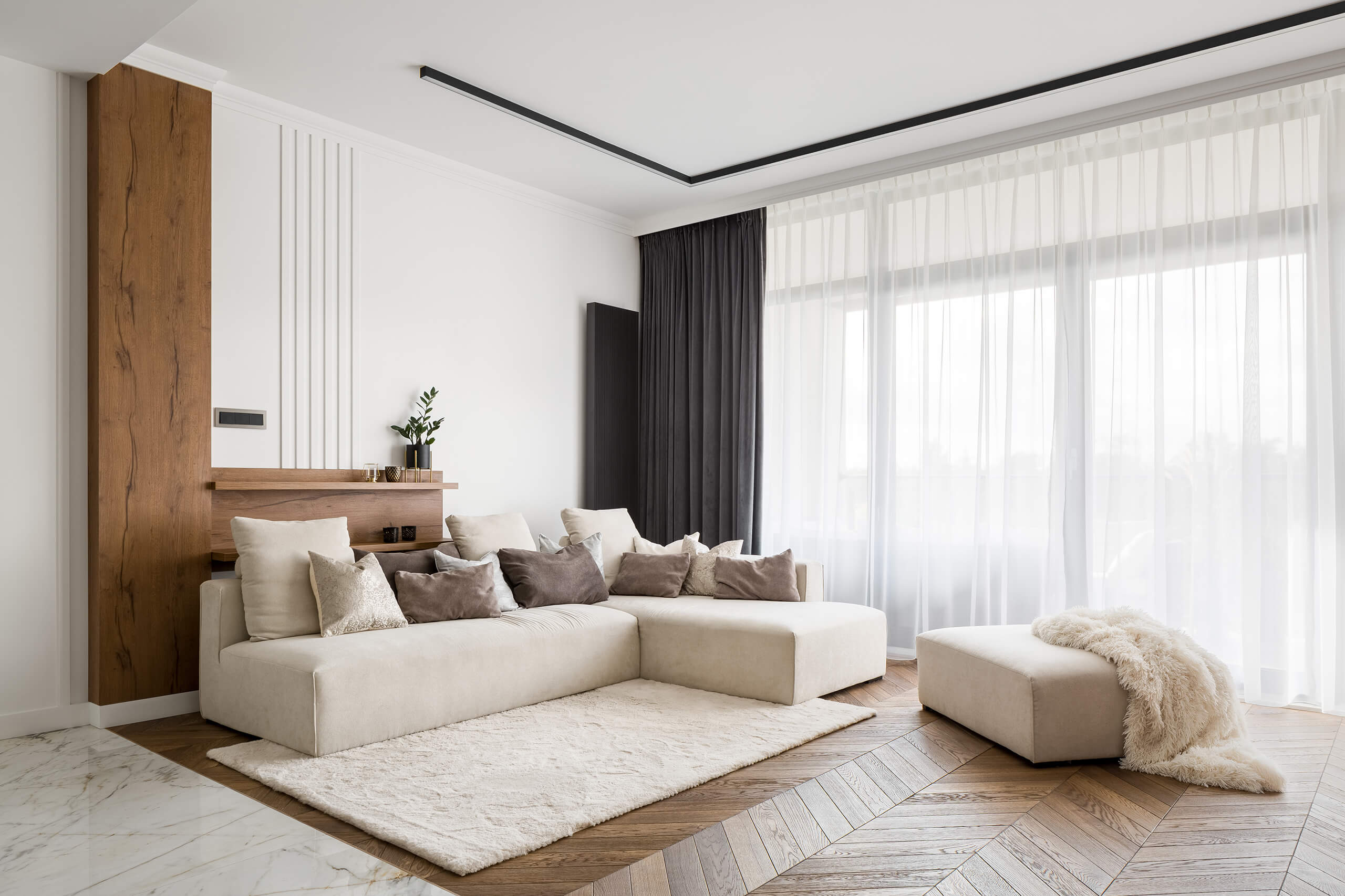 Beige Sofa, braune Kissen, Hocker, großes Fenster mit Gardinen.