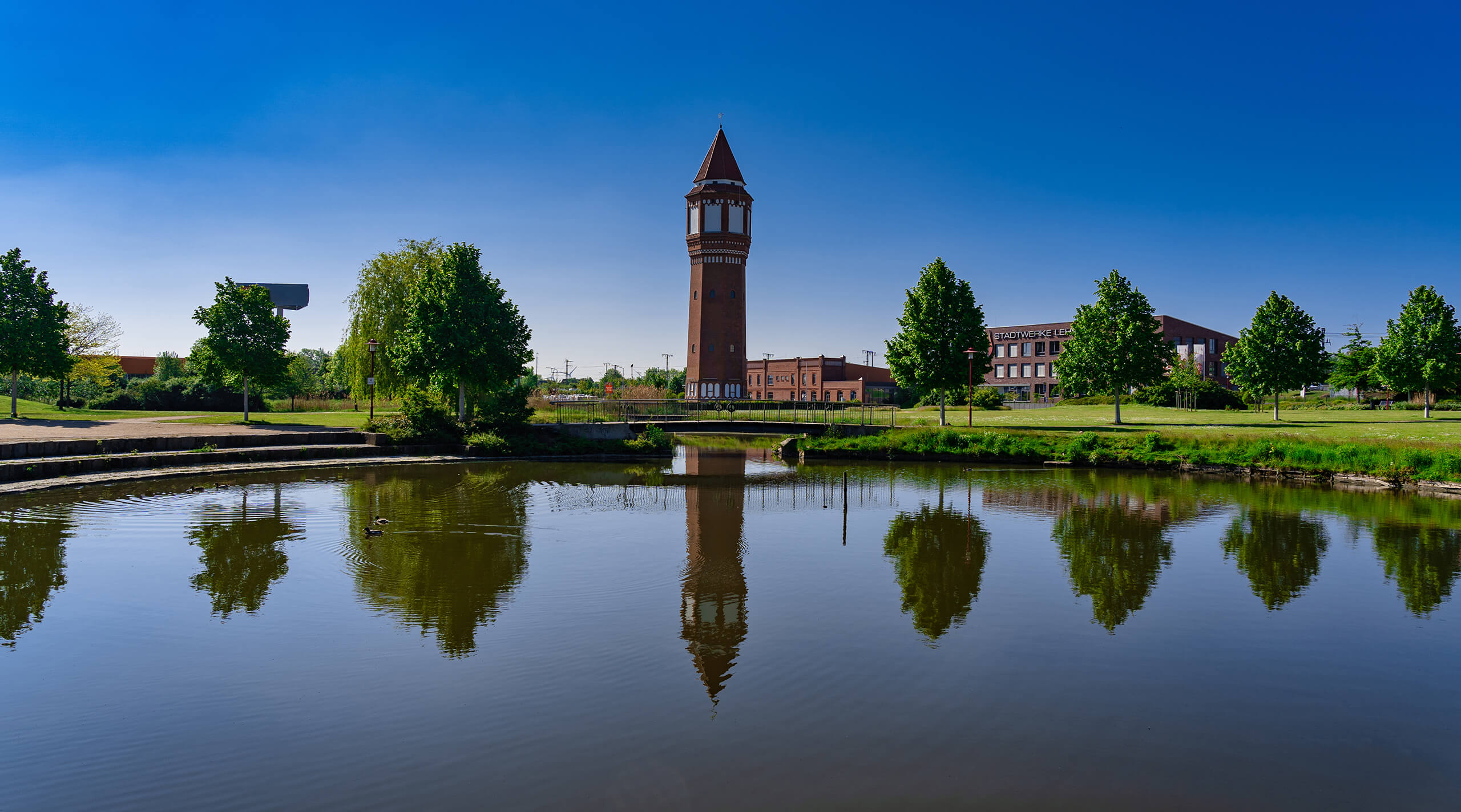 Uhrenturm und Bäume spiegeln sich in einem Teich unter blauem Himmel.